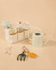 The Gardening Tools Playset includes a beige gardening tote filled with child-friendly tools, a pale watering can, green hand cultivator, orange trowel, and two plant markers—perfect for kids—displayed on a light beige background.