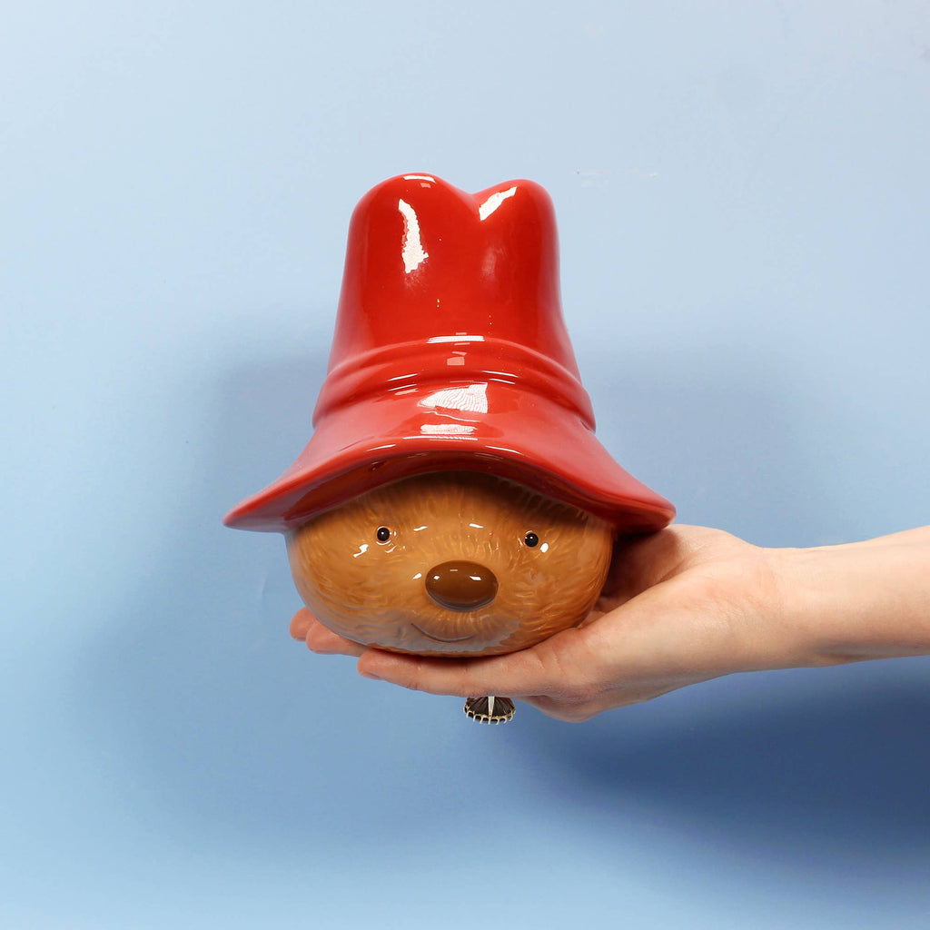 A hand holds the Paddington Money Bank, an officially licensed ceramic coin bank shaped like Paddington Bear’s head with a large red hat, against a plain light blue background.