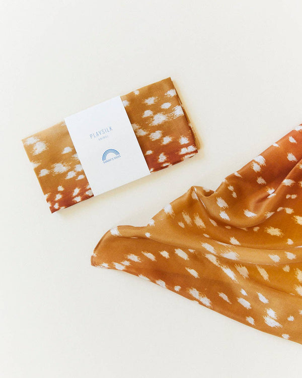A folded silk scarf with a white "PLAYSILK" label lies beside an open Sarah's Silks Animal Print Playsilk - Fawn, featuring white spots on cream—a charming addition to Waldorf or pure silk toy collections.