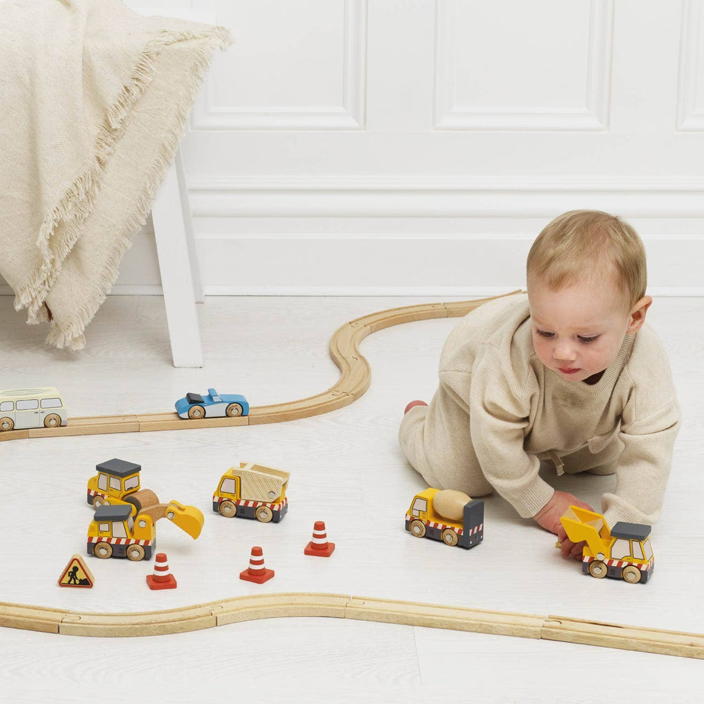 A baby in beige clothes plays on a light floor with the Construction Toy Cars, Trucks & Diggers set (ships in one week), including wooden vehicles, traffic cones, and a curved track. A blanket on a chair nearby encourages imaginative play.