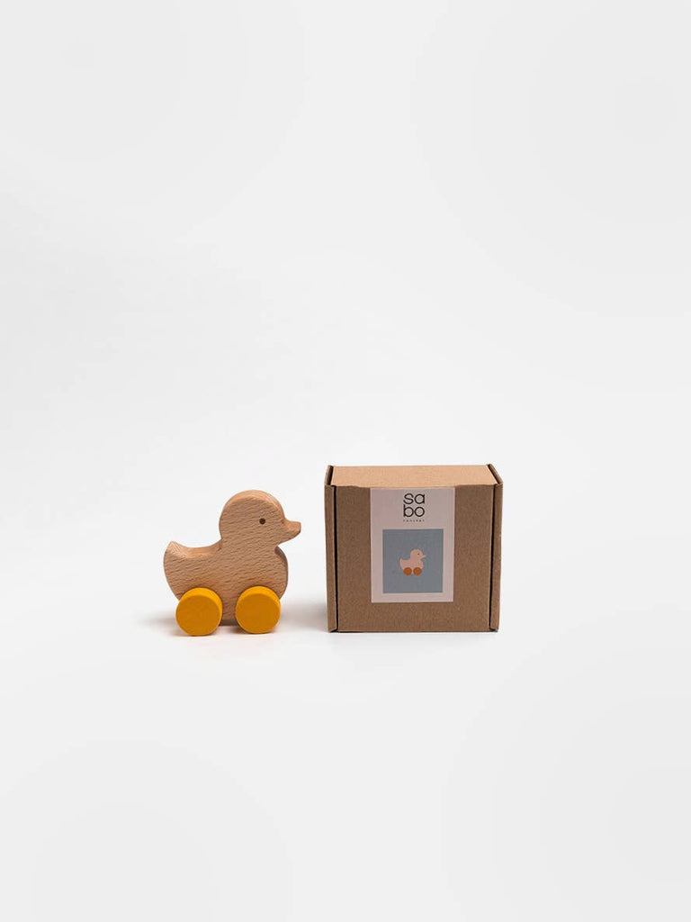 The Sabo Concept Rolling Duck - Yellow, a charming wooden toy with yellow wheels, is placed next to a small brown box featuring its image, both set against a plain white background.