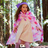 A young child in a dress, lavender leggings, and a purple headband with a star poses playfully in the forest, wearing Sarah's Silks Magical Creatures Playsilk - Pink Unicorn as a cape—perfect for imaginative, open-ended play.