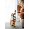 A young girl with blonde hair is playing with the Pound A Ball Tower, a wooden toy featuring colorful discs inside that enhance her motor skills. She is holding a wooden mallet, getting ready to strike the red ball at the top. Dressed in a brown shirt and white pants with a subtle pattern, she enjoys the softly lit background.