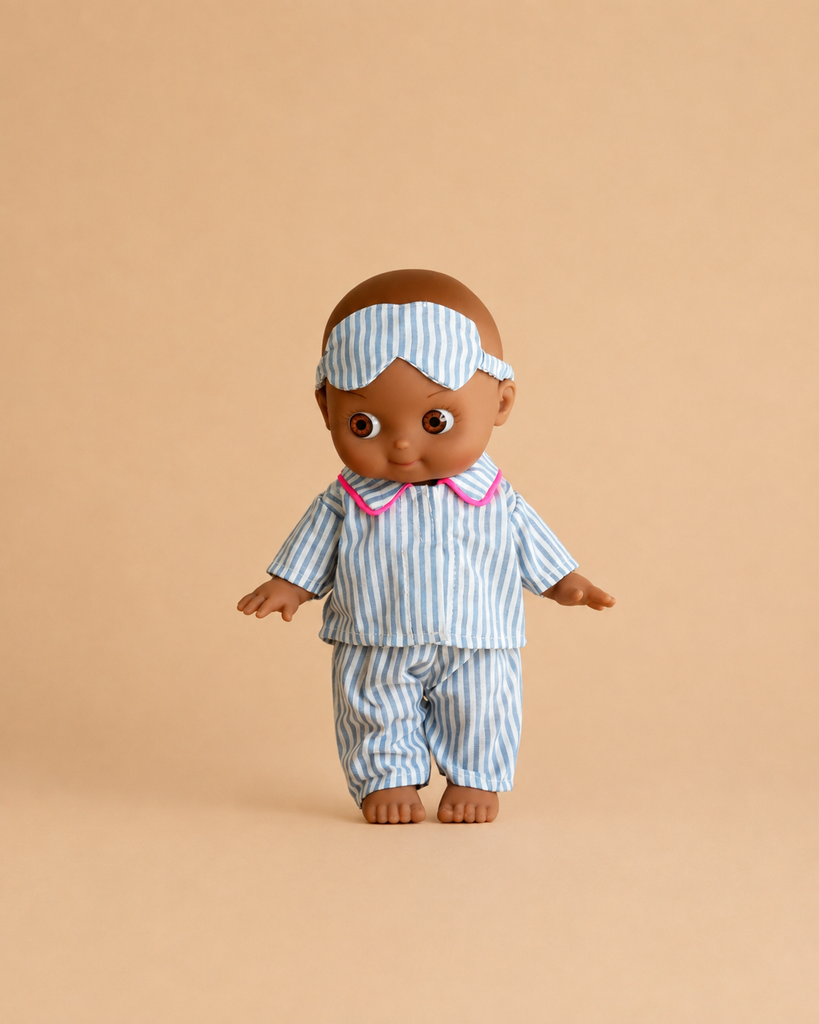 The Minikane Mini Pancha Doll - Noah with Blue Striped Sleepsuit, crafted from high-quality vinyl and featuring blue and white striped pajamas with pink trim and a matching sleep mask, is shown against a plain beige background—perfect for inspiring imaginative play.