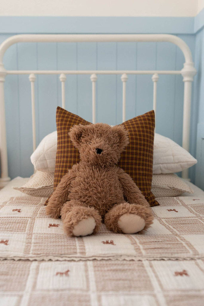 A brown teddy bear sits on a neatly made bed with an Organic Cotton Twin Size Blanket - Pony, a brown plaid pillow, and a white headboard against a light blue wall.