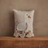 The Coral & Tusk Mama Duck Pocket Pillow, beige with an embroidered white duck, sits against a brown wall. Three removable felt ducklings rest in front on a wooden surface, completing this charming scene.