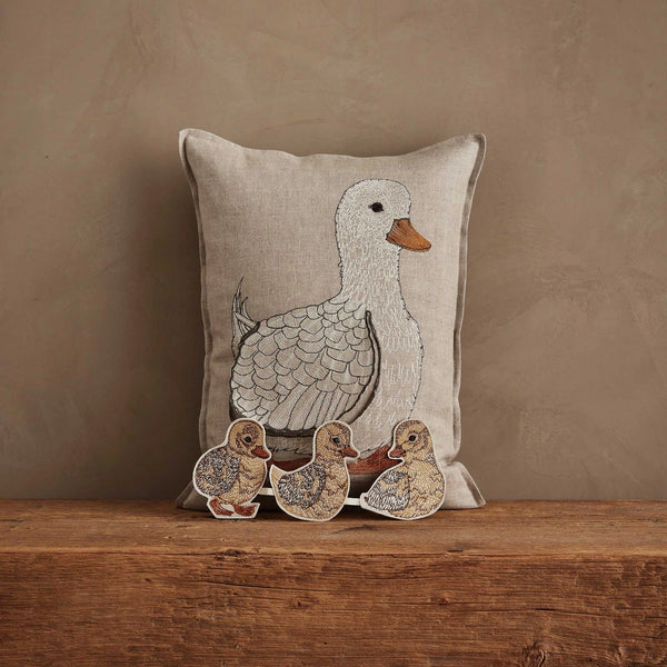 The Coral & Tusk Mama Duck Pocket Pillow, beige with an embroidered white duck, sits against a brown wall. Three removable felt ducklings rest in front on a wooden surface, completing this charming scene.