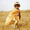 A young child with curly hair wears Sarah's Silks Animal Print Playsilk - Fawn as a cape, along with a matching animal ear headband, yellow leggings, and a skirt, walking through a dry grassy field under a clear sky.