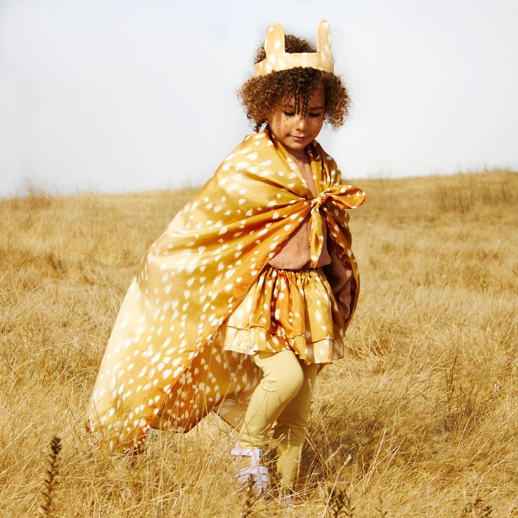 A young child with curly hair wears Sarah's Silks Animal Print Playsilk - Fawn as a cape, along with a matching animal ear headband, yellow leggings, and a skirt, walking through a dry grassy field under a clear sky.