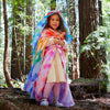 A young child wears the Sarah's Silks Magical Creatures Playsilk - Pink Unicorn, smiling among tall trees in a forest, colorful and ready for imaginative play under the sunlight.