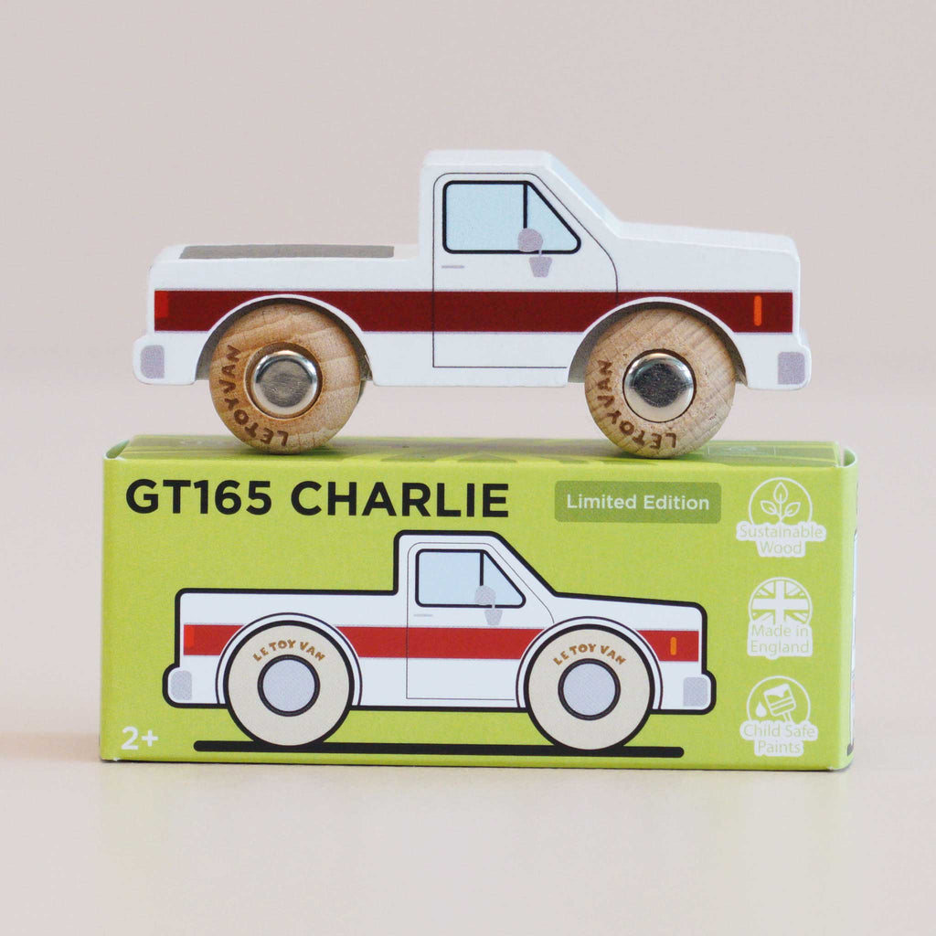 A classic wooden toy truck with red stripes is perched on a green box labeled “No. 30 GT165 Charlie | Made in England.” Crafted from sustainable wood and finished with child-safe paint, this pick-up truck celebrates UK craftsmanship. The box showcases an illustration of the truck, along with icons emphasizing its eco-friendly materials and safety for children.