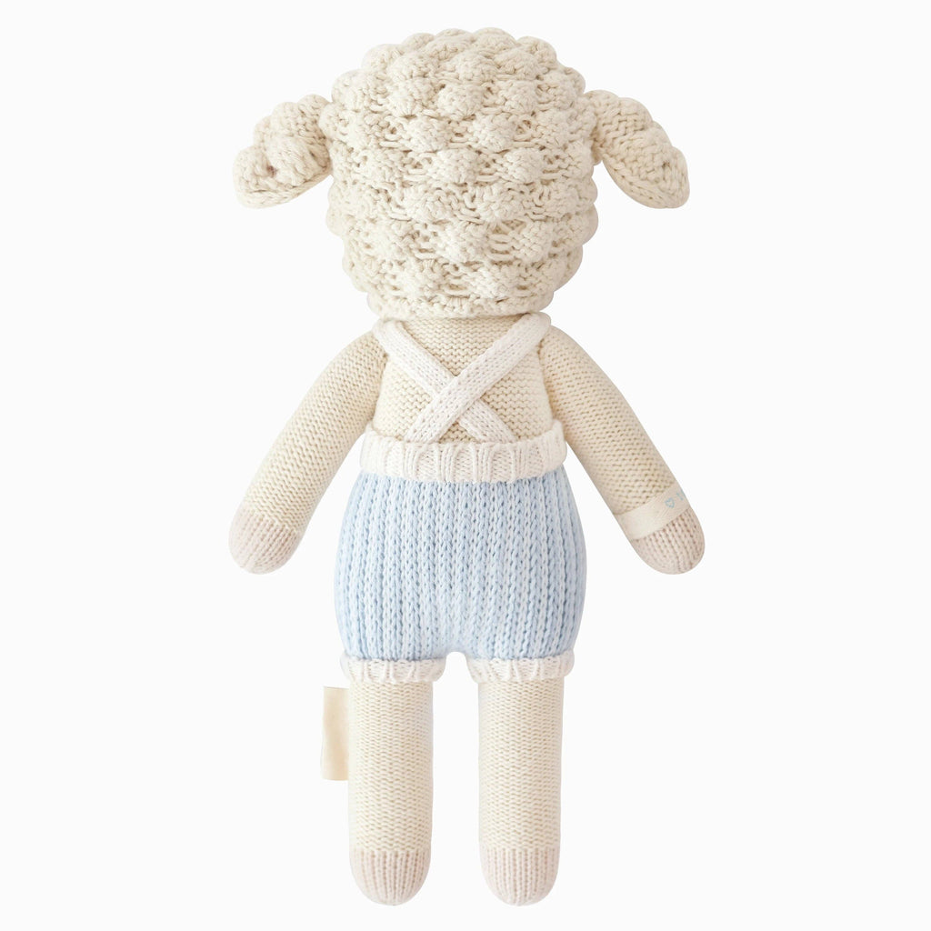 Cuddle + Kind Benjamin the Lamb is a hand-knit plush toy crafted from 100% cotton yarn, featuring a cream textured head with floppy ears, cream body, and light blue overalls with white suspenders. Stands upright on a white background.