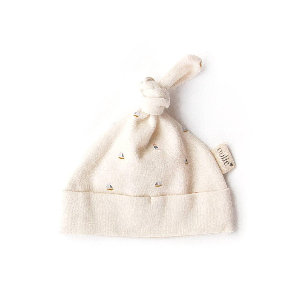 The Organic Baby Hat – Schooner is a soft, cream-colored organic cotton hat featuring a top knot and small sailboat patterns, shown flat on a white background with a side fabric tag—ideal for cozy newborn wear.
