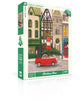 A 1000-piece Christmas Cheer - 1000 Piece Jigsaw Puzzle box featuring a Janet Hill illustration of a red car carrying a Christmas tree in front of storefronts decorated for the holidays.