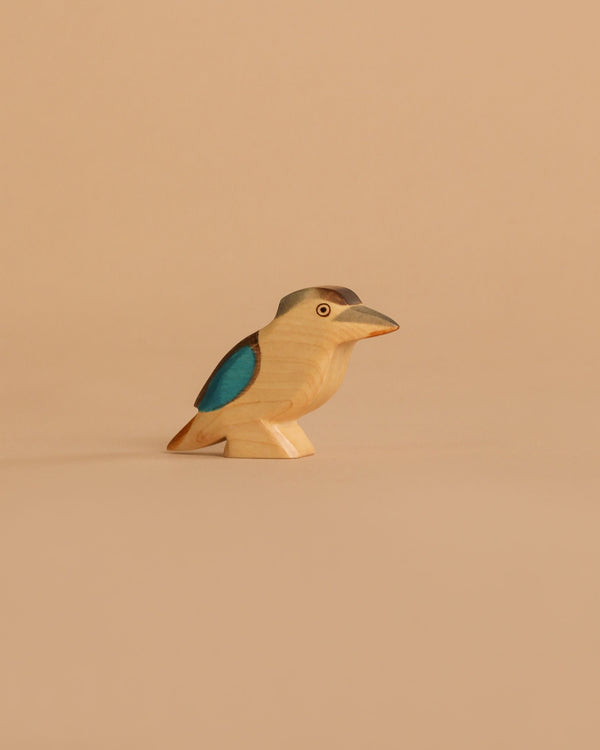 The Handmade Holzwald Kookaburra Bird, featuring blue wings, a gray head, and beige body, stands on a light beige background—a perfect example of beautifully crafted sustainable toys.