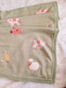 The Organic Cotton Knit Baby Blanket - Farm Animals features soft green fabric with illustrations of pink pigs, a white sheep, yellow ducklings, and a goose. Crafted from organic cotton, it rests on a fluffy white surface.