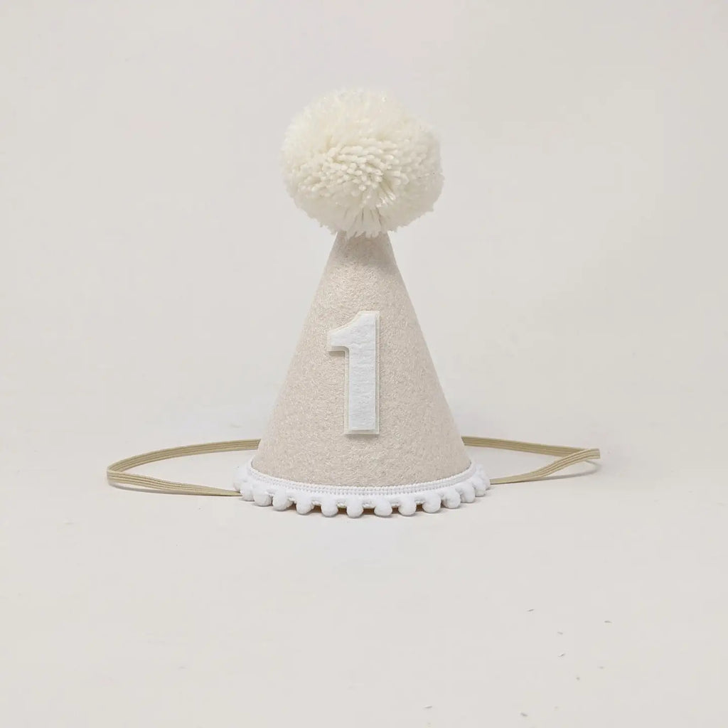 Introducing the White Party Hat: a charming accessory perfect for birthday celebrations. This small, light beige hat features a large white pom-pom on top and is adorned with the number "1" in white on the front. It also boasts a white trim at the base and includes an elastic strap for comfortable wearing. The hat is set against a plain white background, making it an ideal choice for commemorating special occasions.