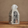 The Coral & Tusk Haunted House Tissue Box Cover, shaped like a house with skeleton and cat illustrations in the window, sits on a wooden surface against a beige wall.