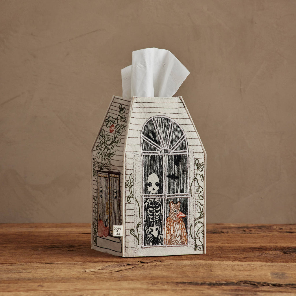 The Coral & Tusk Haunted House Tissue Box Cover, shaped like a house with skeleton and cat illustrations in the window, sits on a wooden surface against a beige wall.