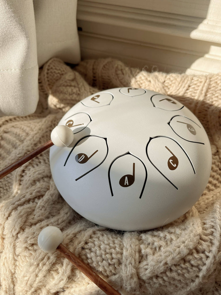 The Steel Tongue Drum, a white musical toy instrument, rests on a knitted beige blanket with two wooden mallets beside it, illuminated by gentle natural sunlight.