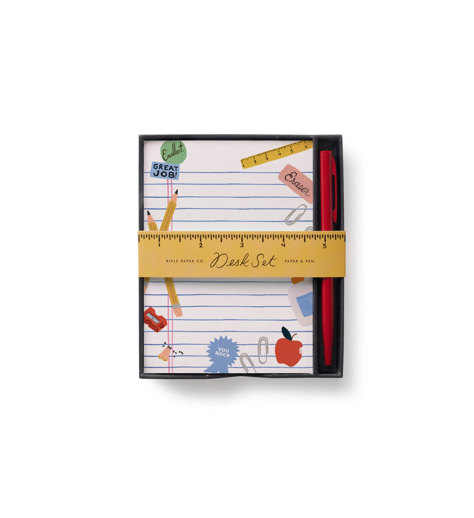 Introducing the Schoolhouse Note Writing Set: a boxed desk set with linen-texture flat notes adorned with school-themed illustrations and paired with a red pen. The reusable desktop box is wrapped in a yellow paper band featuring a ruler design and labeled “Desk Set Paper & Pen.”.