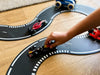 A child pushes a toy car on the Large Flexible Toy Race Track - Grand Prix, curving it alongside other vehicles on a wooden floor. This dynamic race track encourages imaginative play and creative racing fun.
