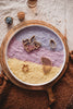 A round tray featuring layered purple and yellow textured powder, adorned with wooden unicorn, cloud, star, and rainbow shapes on a beige cloth, with bowls of beads nearby—perfect for sensory play using the Unicorn Cutter Set - Eco Playdough Tool.
