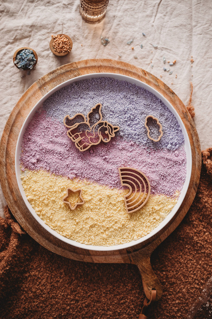 A round tray featuring layered purple and yellow textured powder, adorned with wooden unicorn, cloud, star, and rainbow shapes on a beige cloth, with bowls of beads nearby—perfect for sensory play using the Unicorn Cutter Set - Eco Playdough Tool.