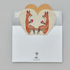 Two Coral & Tusk Smitten Foxes facing each other on a Coral & Tusk Smitten Foxes Felt Envelope, placed on an open envelope with a tree logo, on a grey background.