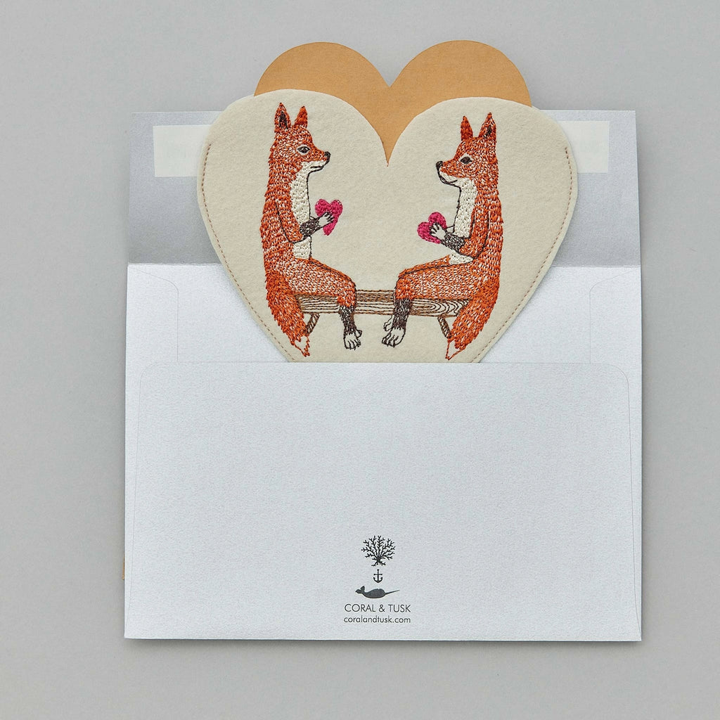 Two Coral & Tusk Smitten Foxes facing each other on a Coral & Tusk Smitten Foxes Felt Envelope, placed on an open envelope with a tree logo, on a grey background.