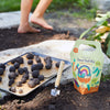 A barefoot person stands in a garden beside soil, a trowel, a tray of seed balls, and the colorful DIY Seed Ball Kit - Rainbow Garden box—a fun STEM activity with Non-GMO Seeds and illustrated veggies and rainbow on the packaging.