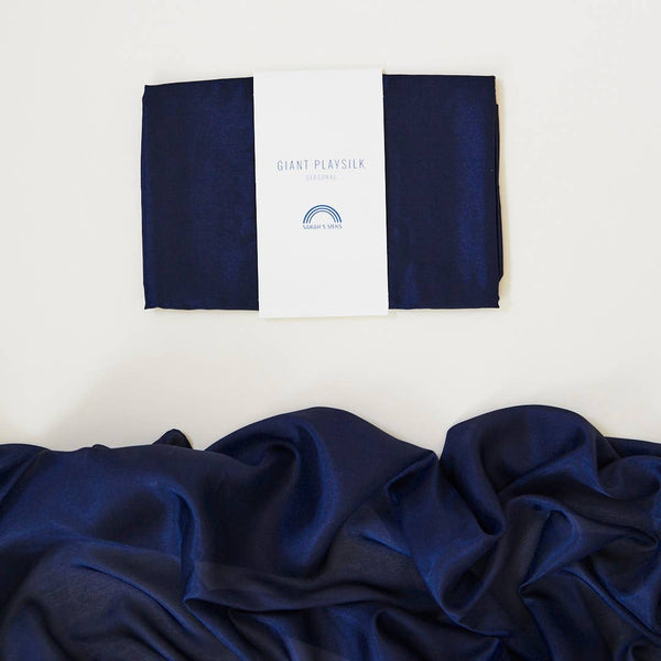 A neatly folded navy blue playsilk is displayed with a white label reading "Sarah's Silk Giant Midnight Playsilk." Below, the same fabric is spread out in soft, flowing folds, inviting imaginative play and showcasing its smooth and shiny texture against a light background.