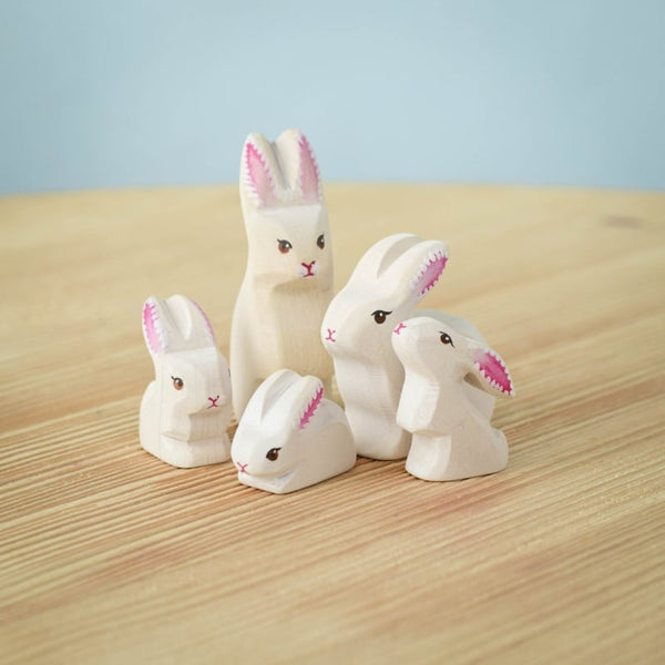 The Bumbu Handmade Wooden White Bunny Rabbit Family Set features five eco-friendly wooden bunnies with pink ears and black eyes, arranged on a light wood surface against a soft blue background—ideal for Montessori toy collections.