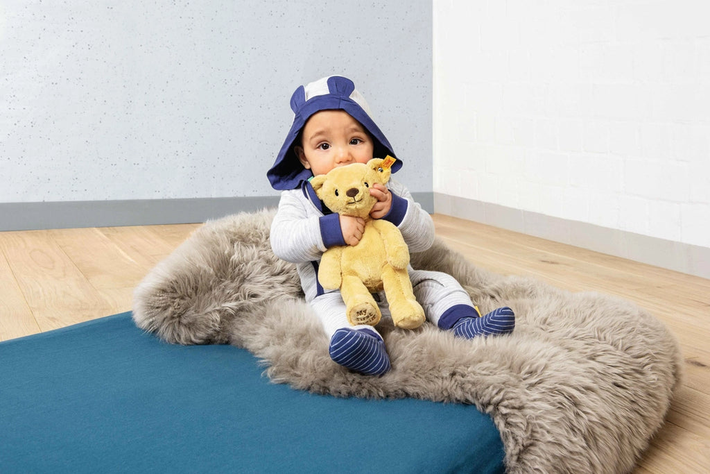 A baby dressed in a cozy animal-themed hoodie and socks sits on a fluffy rug, happily holding a yellow "My First Steiff" teddy bear. The background is minimal with a plain wall and Steiff, My First Steiff Teddy Bear, Beige, 10".