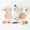 A flat lay displays the Wooden Tea Set & Tray, which includes a charming eco-friendly wooden teapot, three mugs, and two saucers with elegant floral designs. The set is completed with two heart-shaped cookies, a wooden tea bag, and a teaspoon, offering endless possibilities for imaginative play on the pristine white surface.