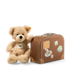 A Steiff, Fynn Teddy Bear in Suitcase next to a vintage-style small suitcase decorated with travel stickers, against a plain, light background.