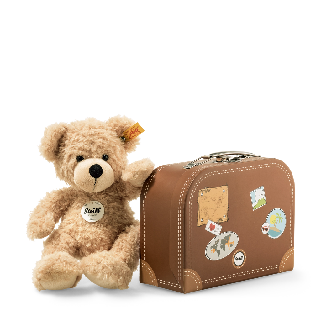 A Steiff, Fynn Teddy Bear in Suitcase next to a vintage-style small suitcase decorated with travel stickers, against a plain, light background.