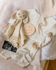 A flat lay of baby items features the Cuddle + Kind Organic Cotton Baby Bunny Lovey (Oatmeal), cream baby clothes, a wooden "hello world" sign, baby socks, and an ultrasound photo on a soft fabric background.