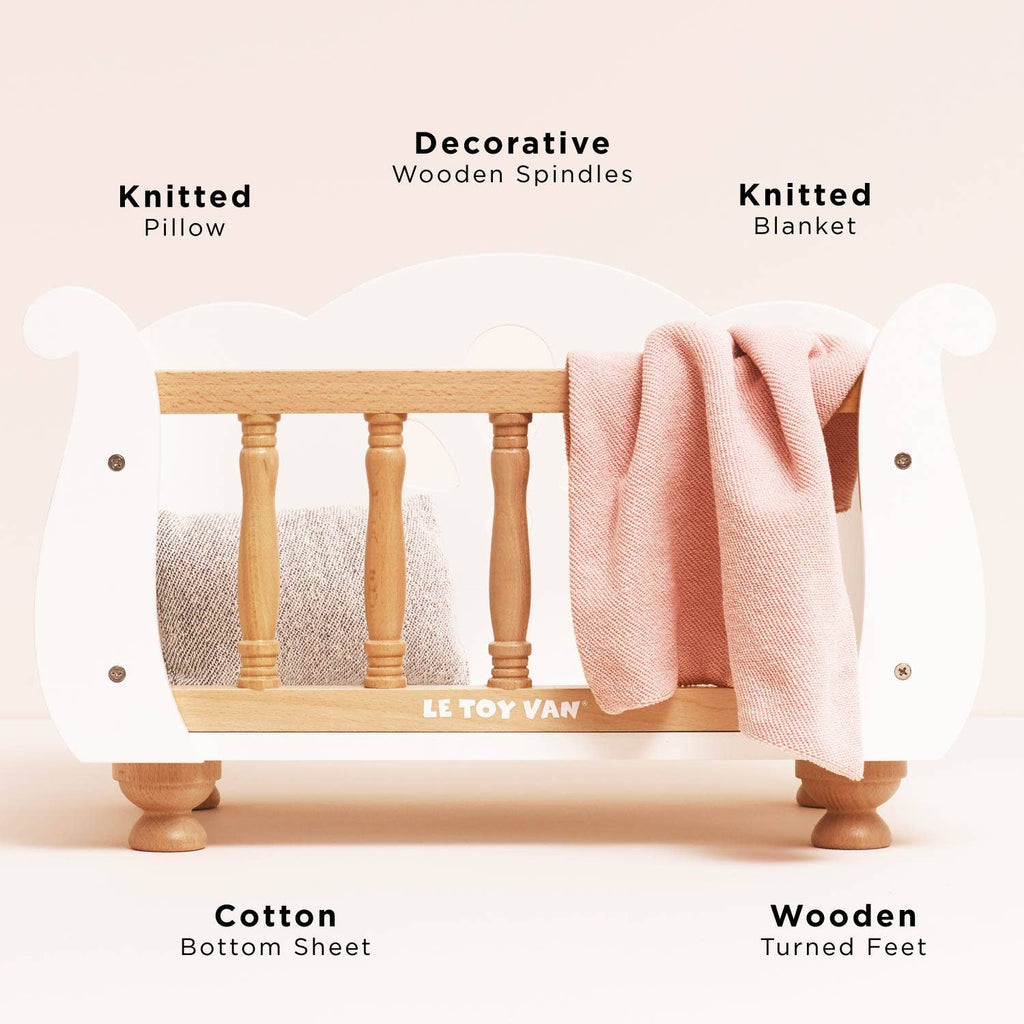 A Wooden Dolls Cot with turned feet and a soft bedding set, featuring labels pointing to the knitted pillow, spindles, blanket, sheet, and feet.