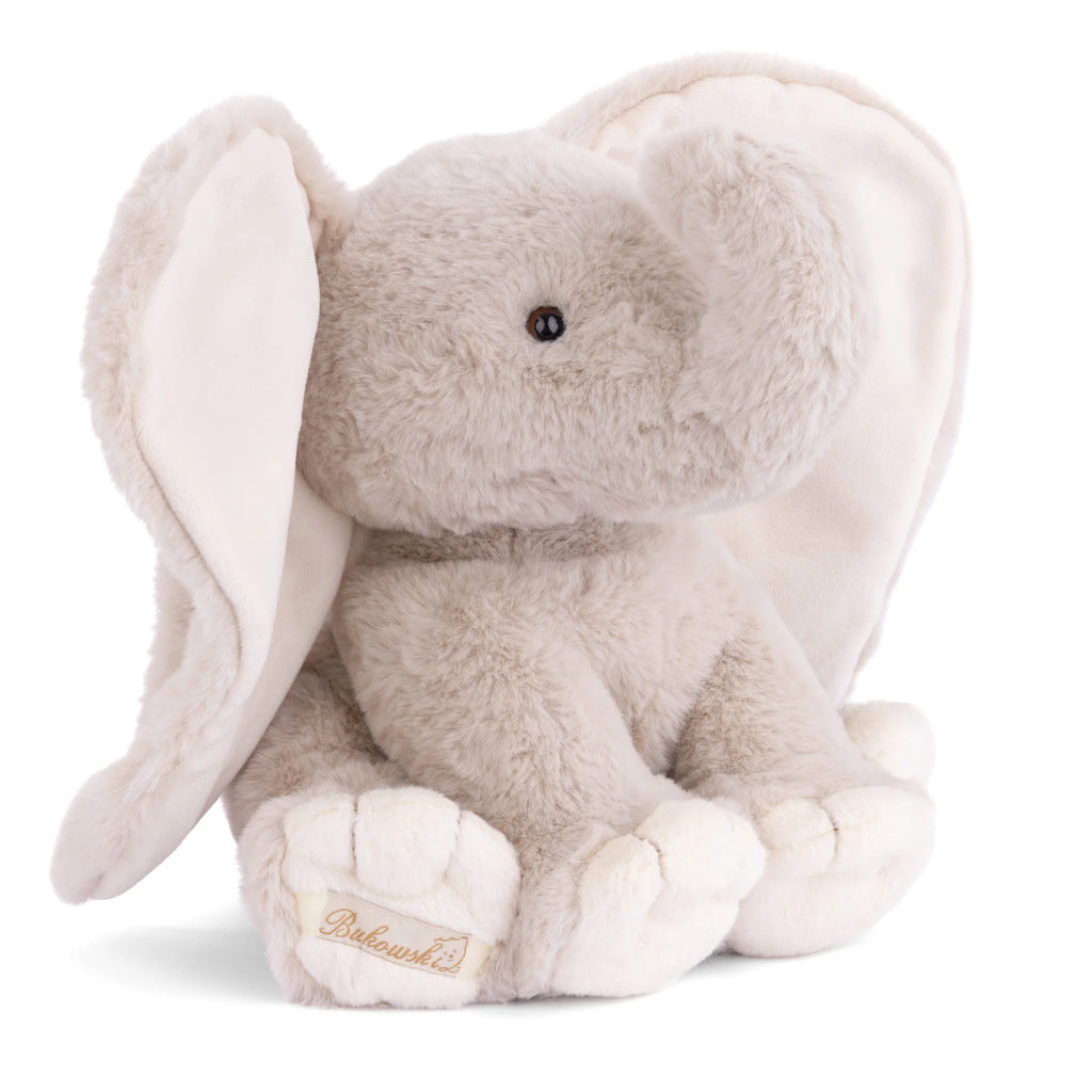 A soft, plush Bukowski - August Elephant toy, featuring floppy ears, light gray fur, and cream-colored accents on its feet and inner ears, sits upright. The brand name "Bababushka" is embroidered on its foot. This huggable elephant makes the perfect bedtime snuggle plush for children of all ages.