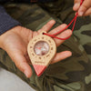 A child holds the Huckleberry Compass, made from FSC-certified beechwood with cardinal directions marked, hanging on a red string—perfect for outdoor adventures. The child wears green camo pants and a dark long-sleeve shirt.