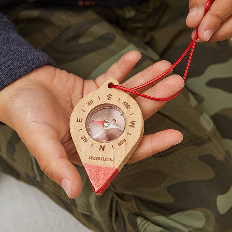 A child holds the Huckleberry Compass, made from FSC-certified beechwood with cardinal directions marked, hanging on a red string—perfect for outdoor adventures. The child wears green camo pants and a dark long-sleeve shirt.