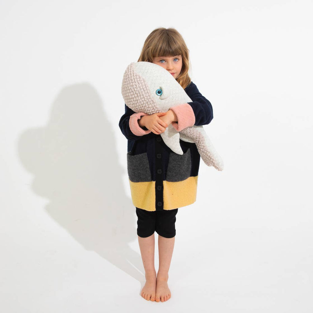 A young child with blonde hair stands barefoot against a white background, cuddling a BigStuffed Small Grandpa Whale. The child is wearing a black, gray, and yellow color-blocked cardigan over black leggings.