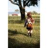 A young child in a pink cap and backpack walks through a grassy field toward a tree, with the Senger Naturwelt Stuffed Animal - Small Sheep peeking out. Sunlight creates soft shadows, and a building appears in the background.
