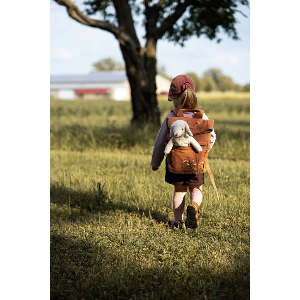 A young child in a pink cap and backpack walks through a grassy field toward a tree, with the Senger Naturwelt Stuffed Animal - Small Sheep peeking out. Sunlight creates soft shadows, and a building appears in the background.