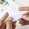A person holds a small white paper with a 4x4 grid of numbers, labeled "Paint By Number Kit - New York" at the top—a compact kit that ignites creativity. Another hand gently touches the corner of this miniature artwork.