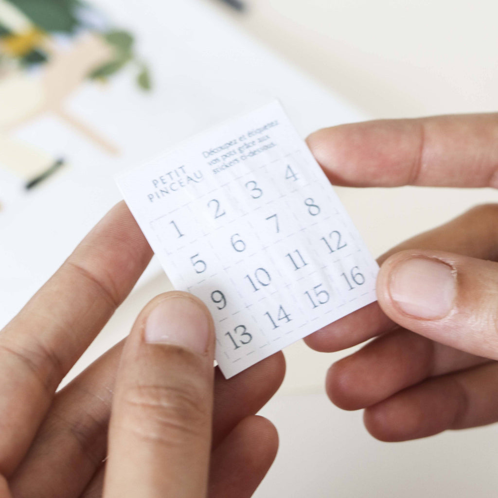 A person holds a small white paper with a 4x4 grid of numbers, labeled "Paint By Number Kit - New York" at the top—a compact kit that ignites creativity. Another hand gently touches the corner of this miniature artwork.
