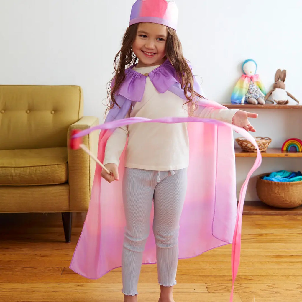 A young girl with curly hair, wearing a paper crown and a flowing pink cape, joyfully engages in open-ended play with a Sarah's Silk Large Heart Streamer Wand in a cozy living room setting.
