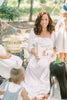 A woman in a white dress holds Winnie-the-Pooh: Finding Feelings while smiling at children in party hats during an outdoor picnic with bunting and a cake on a tree stump.