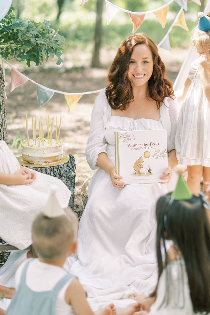 A woman in a white dress holds Winnie-the-Pooh: Finding Feelings while smiling at children in party hats during an outdoor picnic with bunting and a cake on a tree stump.